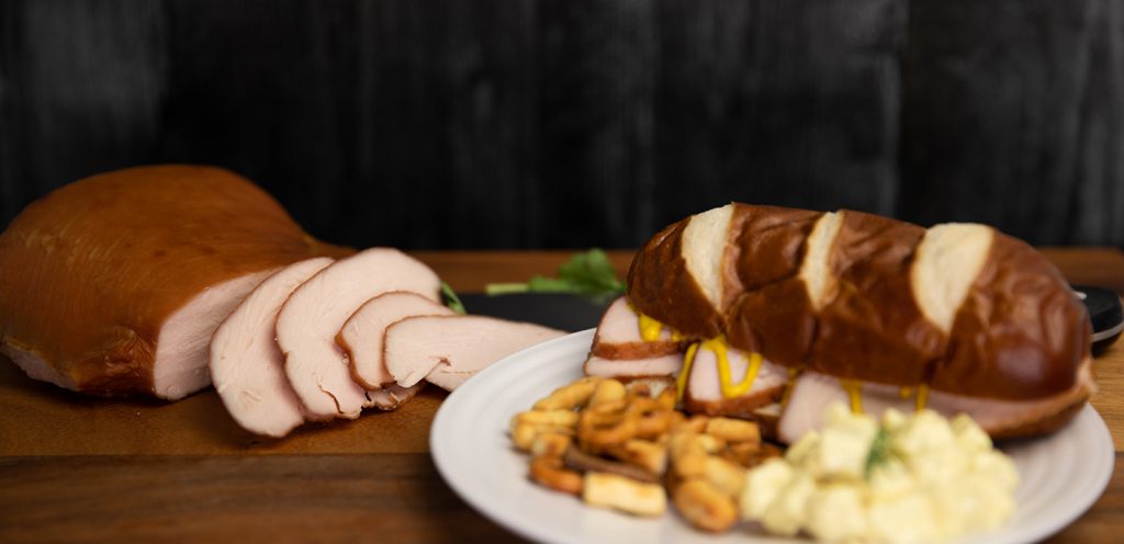 Banner image of the Trig's Smokehouse Smoked Turkey Breast sliced and shown served on a pretzel bun in a sandwich.